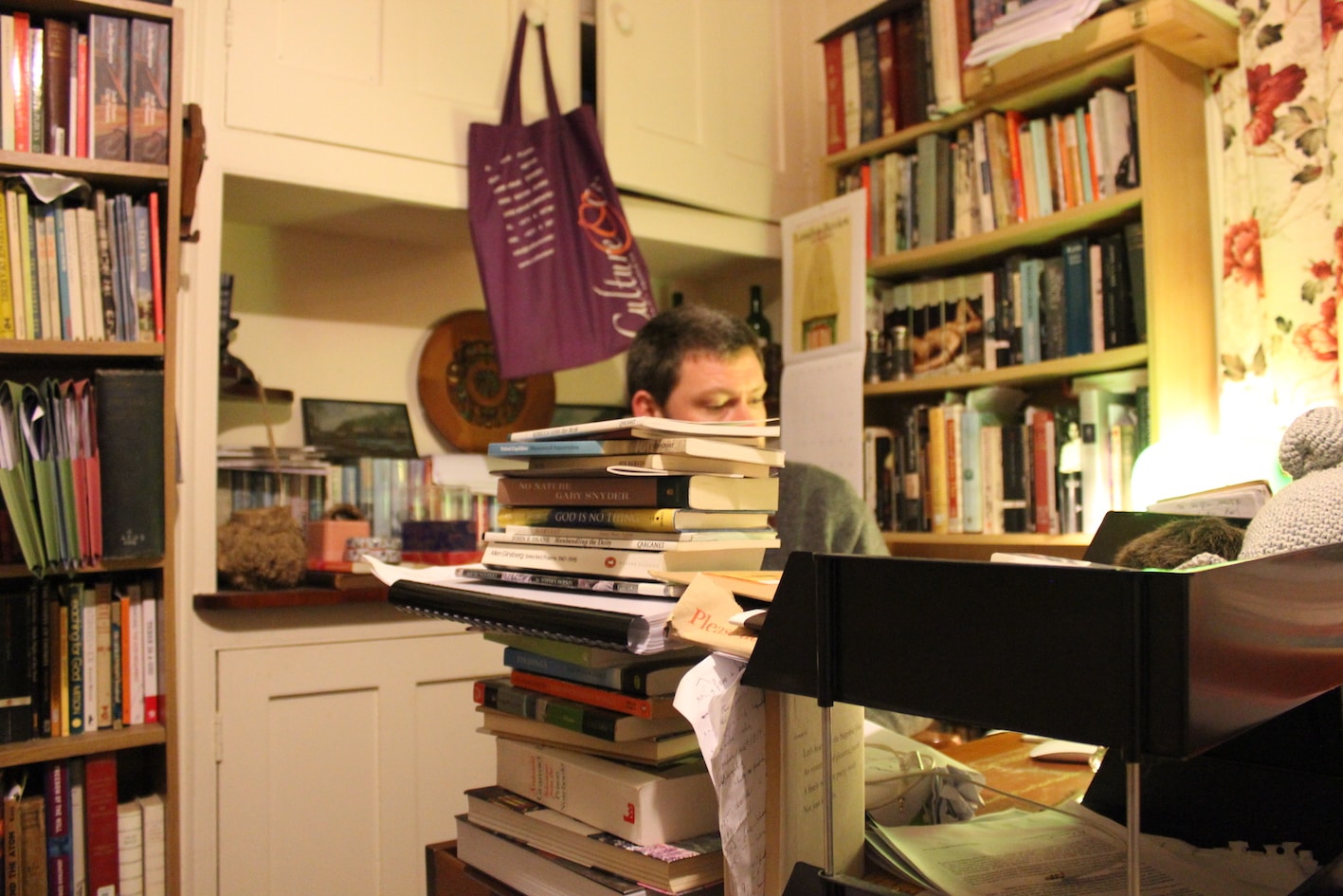 Full colour picture of the editor at his desk though the editor is hidden behind a tower of books. You can see his eyes (looking at something below, perhaps a book or screen, and the top of his head. There are books EVERYWHERE.