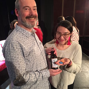 Photo (full colour) shows Stuart Bartholomew and Cynthia Miller beaming delightedly at the camera. Stuart is holding a bottle of beer with what looks like a Verve design label. He is wearing a long sleeved, patterned cotton shirt and has a huge smile and a slightly greying beard -- perhaps early to mid forties. Cynthia is small, much smaller and has gleaming dark hair tied back and huge framed glasses and what could be giant pink earrings. She has something in her hand -- looks like a blue button or badge.